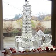 Clear glass lighthouse tower centerpiece on a windowsill, decorated with red rose petals and a heart-shaped ornament.
