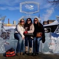 Three people in winter clothes sitting outdoors on a snowy bench under a blue sign.
