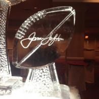 Clear glass football-shaped award with engraved signature on a stand, lit warmly in a dim room