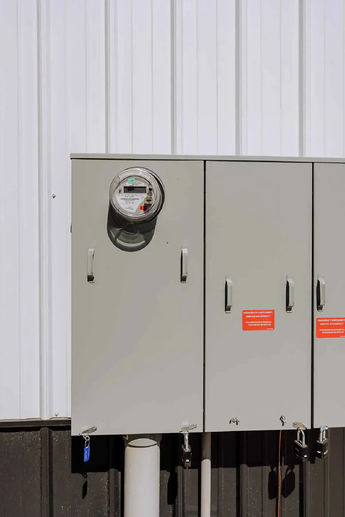 An electric meter attached to a gray, multi-section electrical panel box mounted on a white corrugated metal wall.