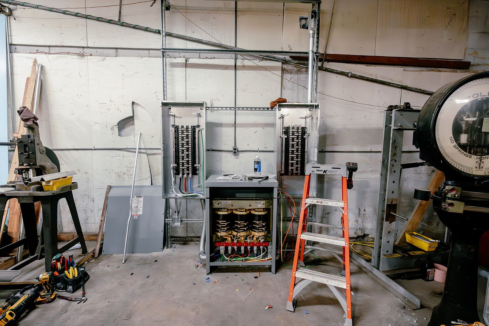 A wall-mounted electrical panel installation under construction in a workshop, featuring conduits and a step ladder.