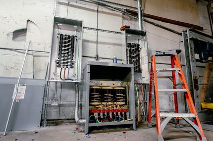 Two open electrical panel boxes flanking a transformer on a wall in an industrial setting, with an orange stepladder nearby.