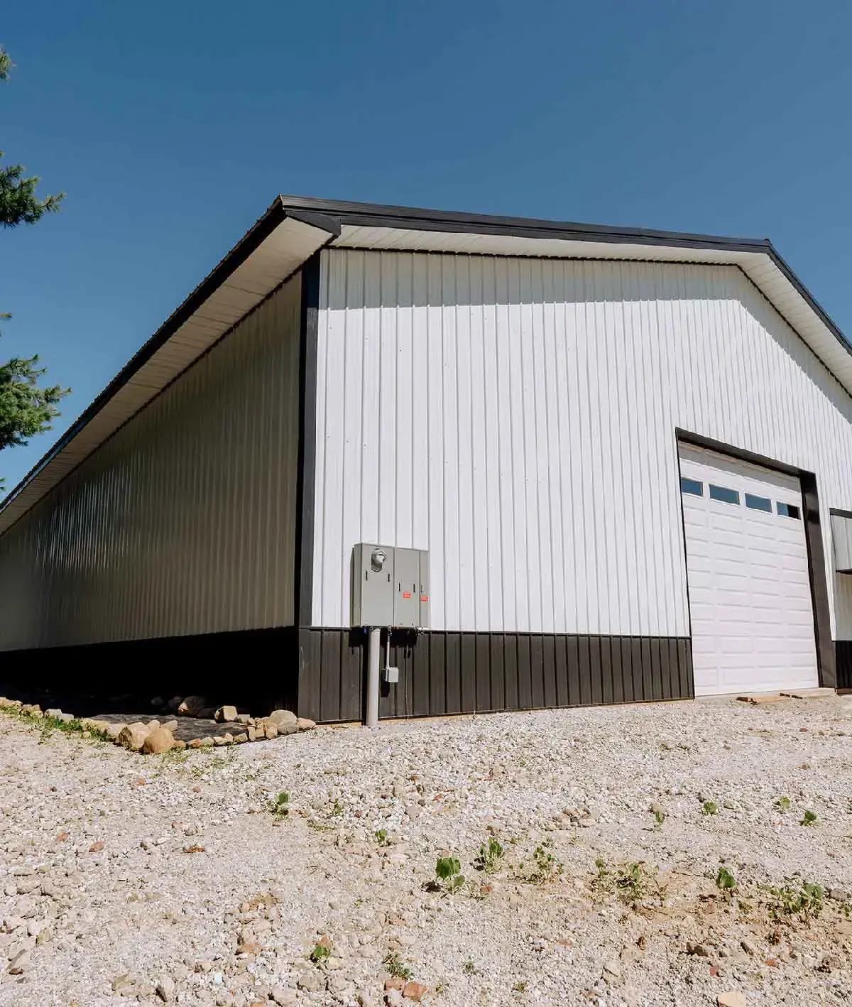 A white metal garage with a black trim and base, a roll-up door, and an electrical box, set on a gravel lot under blue sky.