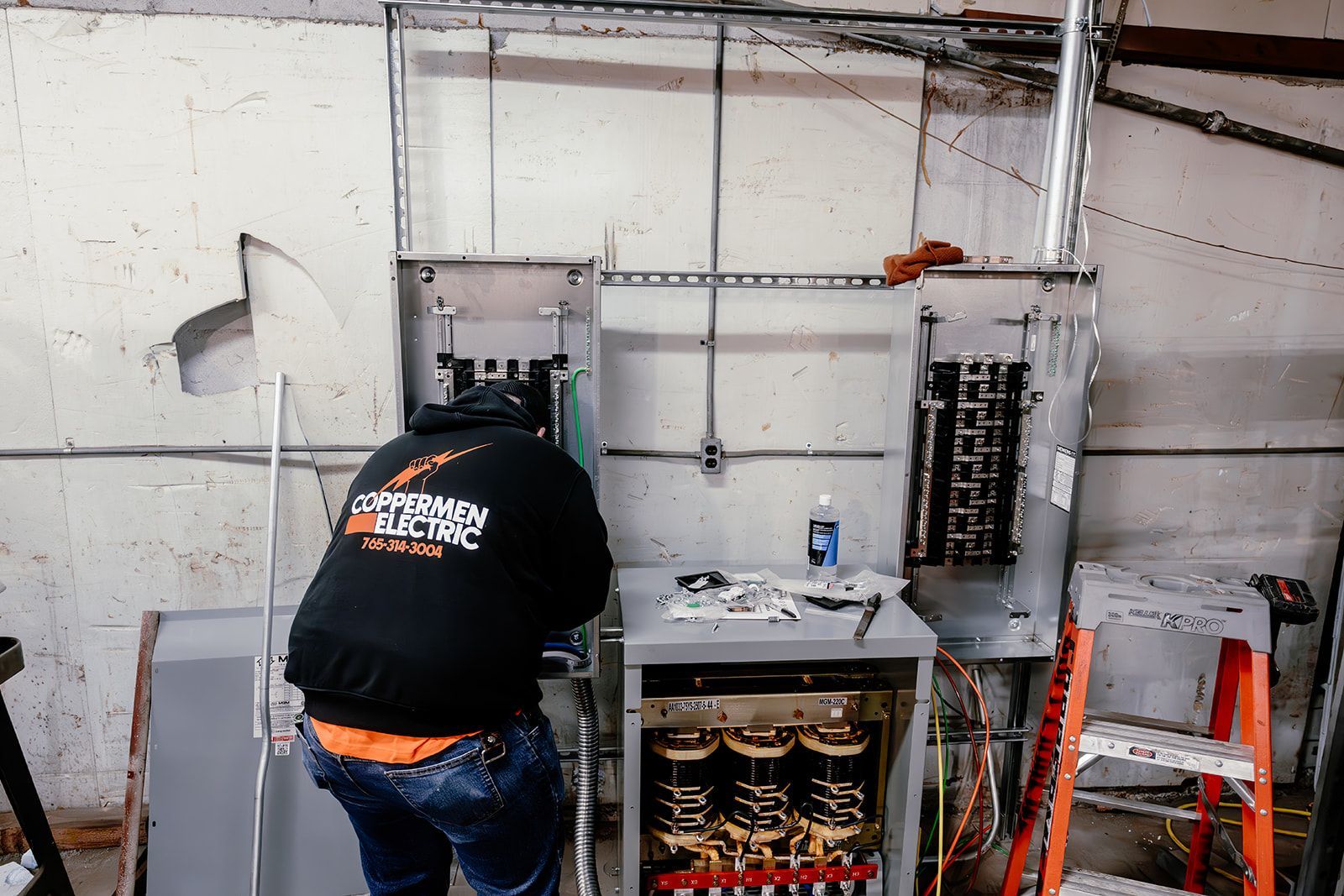 A person wearing a dark hoodie works on exposed electrical panels mounted on a gray industrial wall.