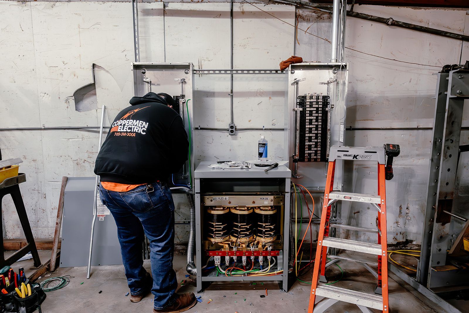 An electrician works on an open electrical panel mounted on a concrete wall next to a step ladder in a workshop.