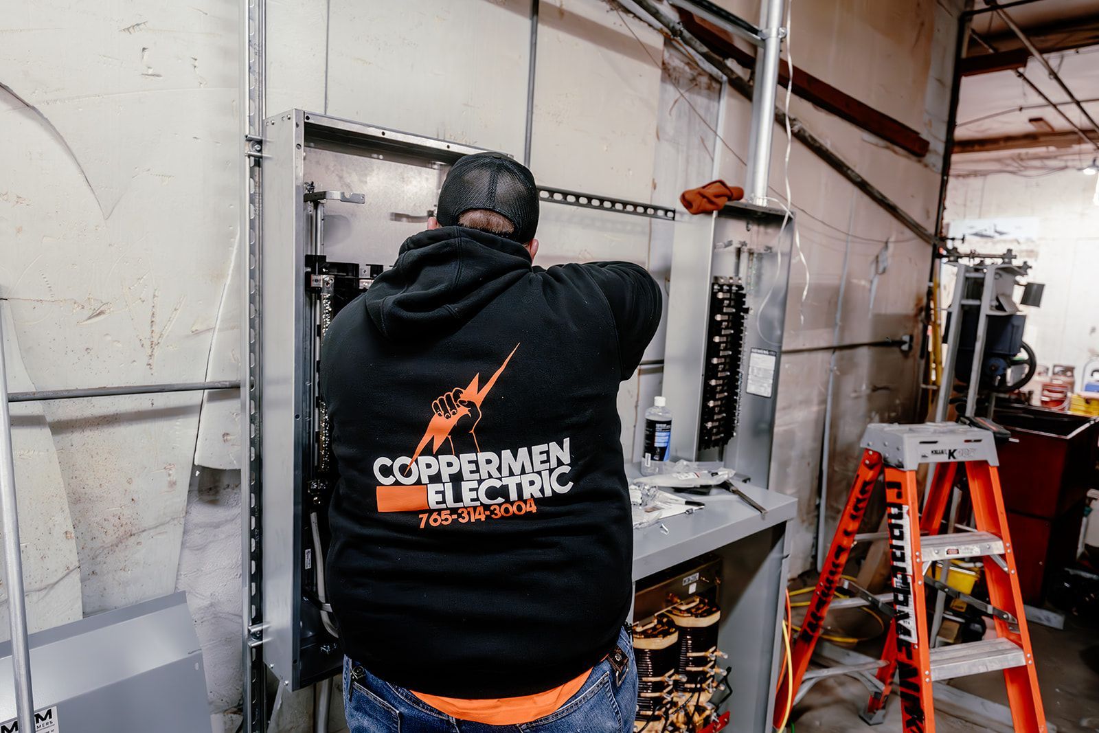 An electrician in a black Coppermen Electric hoodie working on an open electrical panel in an industrial setting.