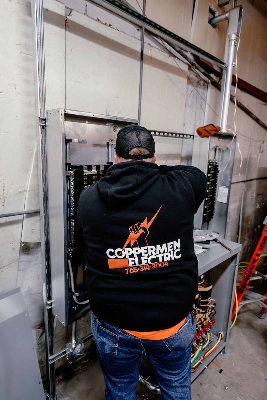 An electrician in a black hoodie works on an open electrical panel mounted to a wall.