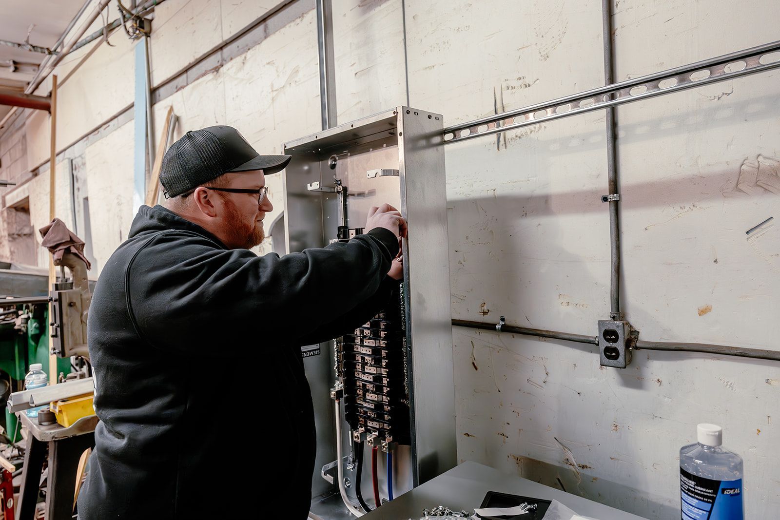 A technician in a black hoodie and cap works on the wiring of an open industrial electrical panel mounted on a wall.