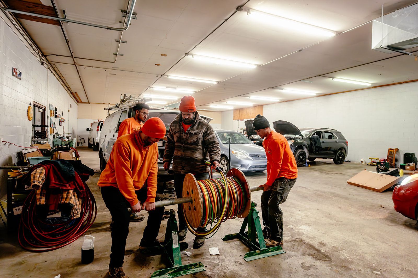 Three workers in orange high-visibility clothing unwind a large industrial cable reel inside a bright, spacious garage.