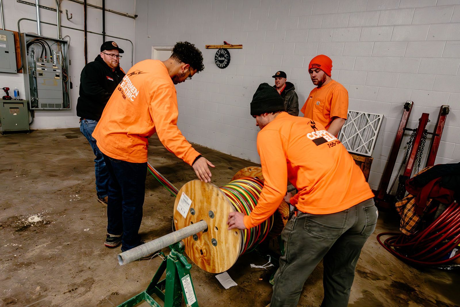Four people wearing orange shirts work with a large cable reel on a stand inside a utility room with concrete walls.
