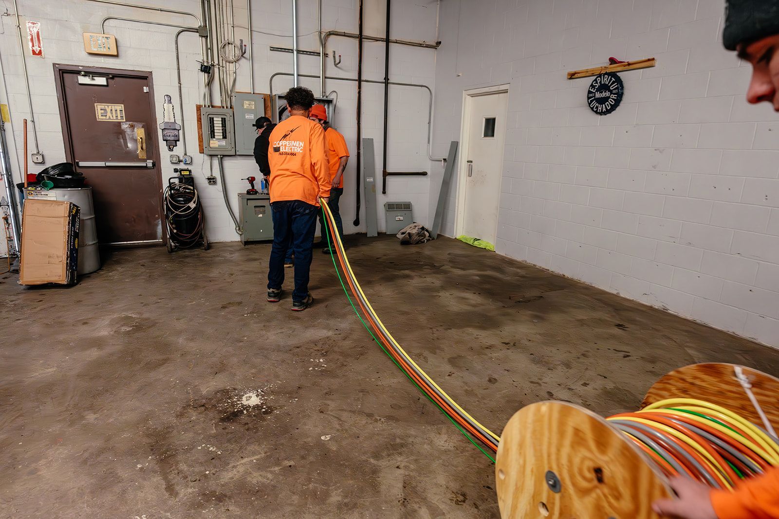 Workers in orange jackets unspool a long, multi-colored cable from a wooden reel in an industrial room.
