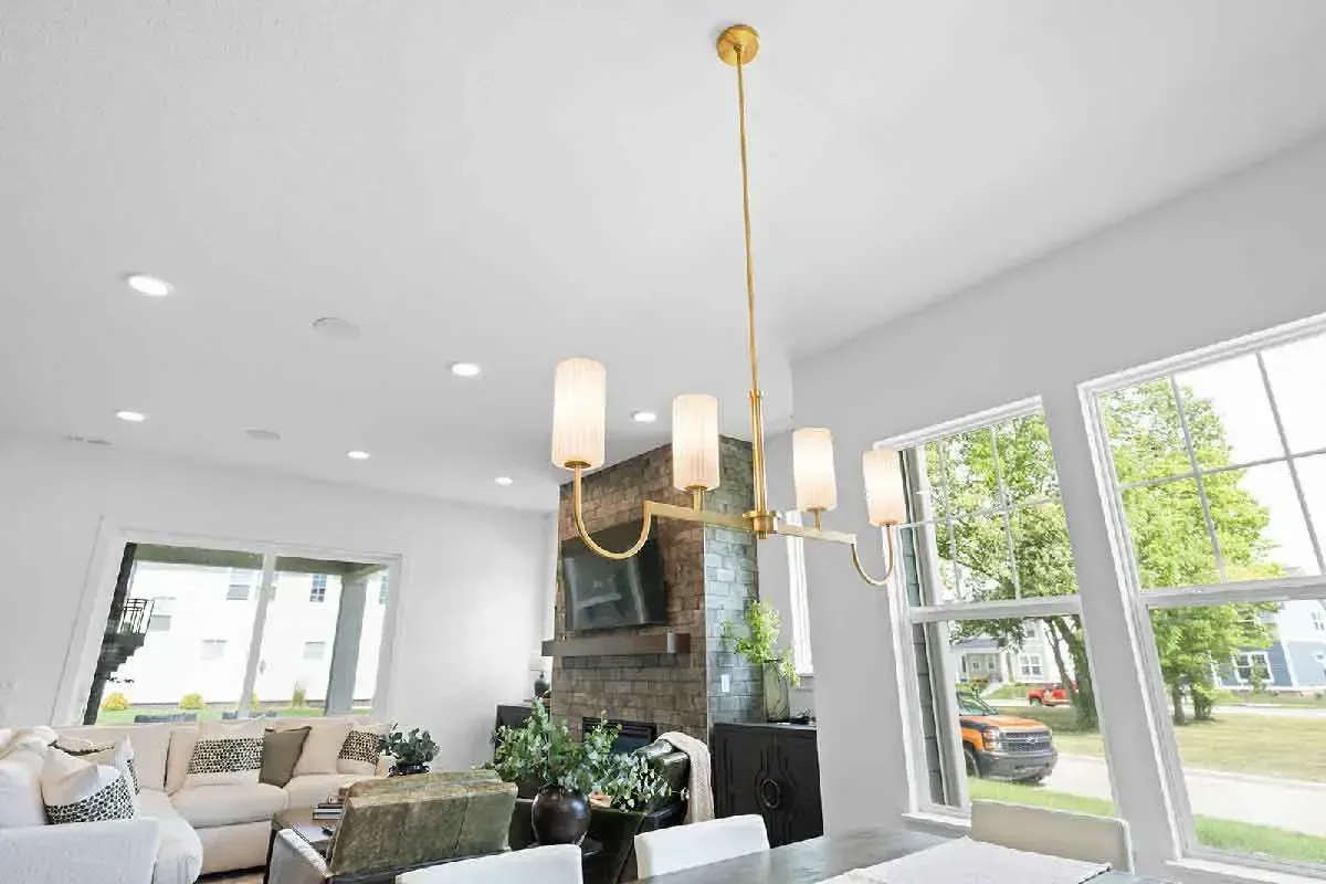 A bright dining room features a modern gold chandelier over a table, with a stone fireplace and windows in the background.