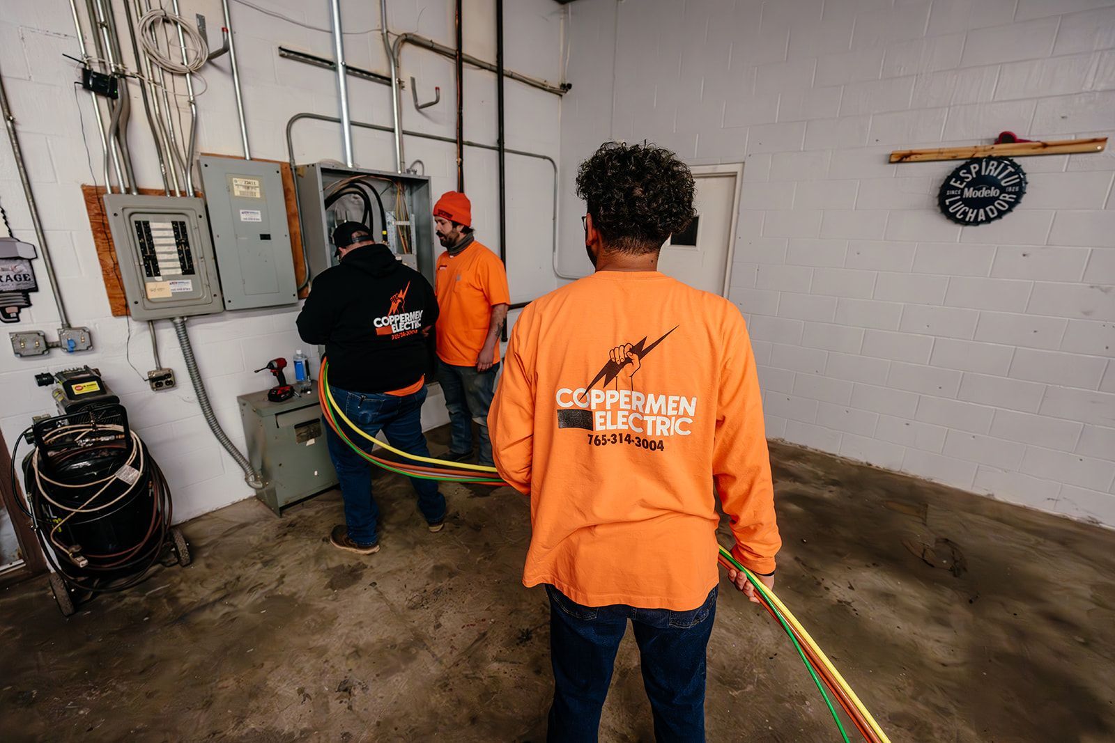 Three workers in high-visibility orange shirts install electrical wiring into a wall-mounted panel in an industrial space.