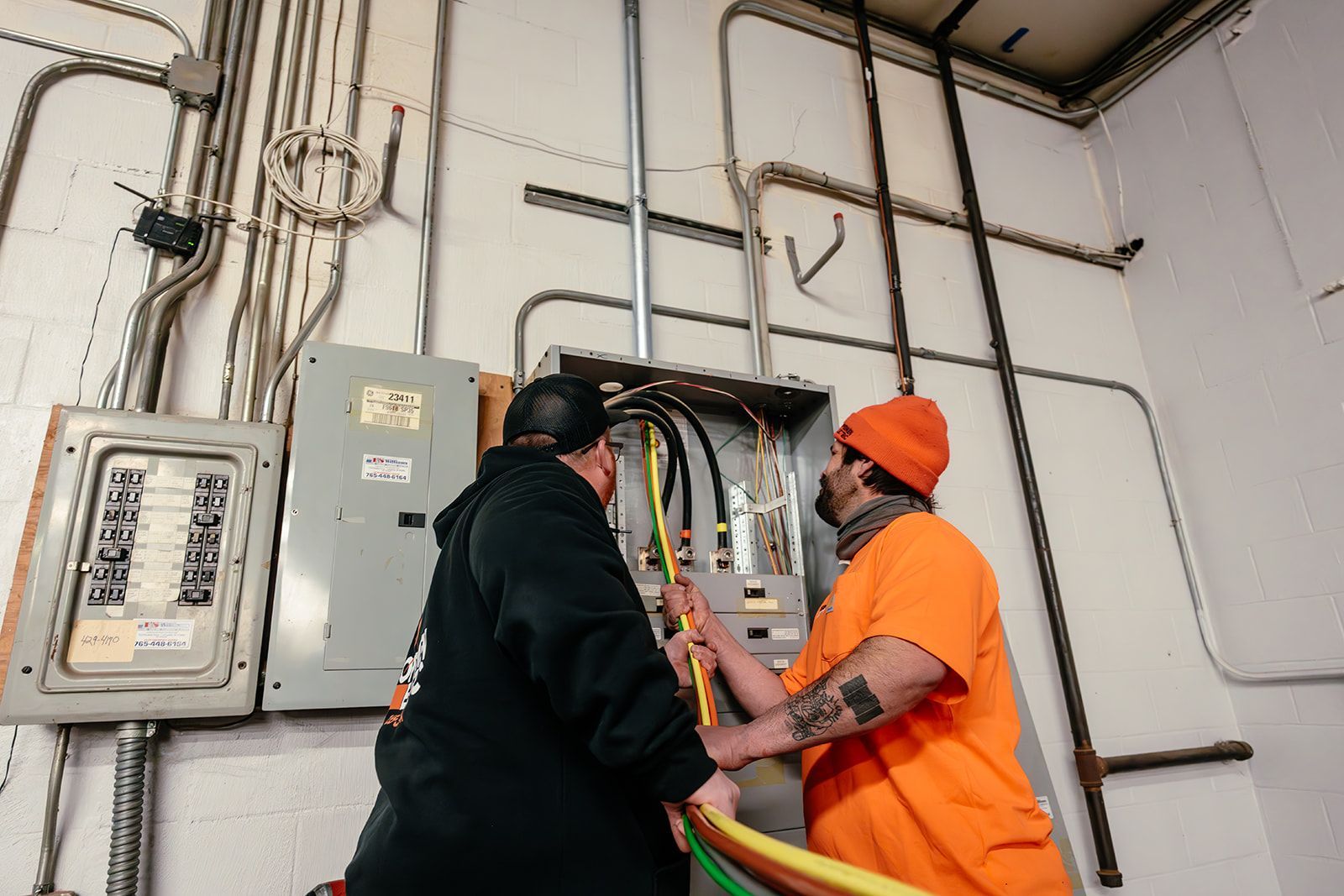 Two workers in a utility room feed thick electrical cables into an open electrical panel mounted on a wall.