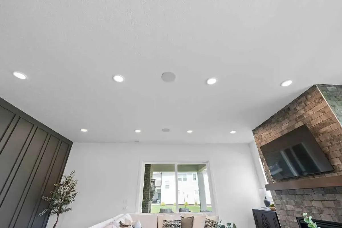 A bright room with a high white ceiling, recessed lighting, a stone fireplace with a TV, and a window overlooking a yard.