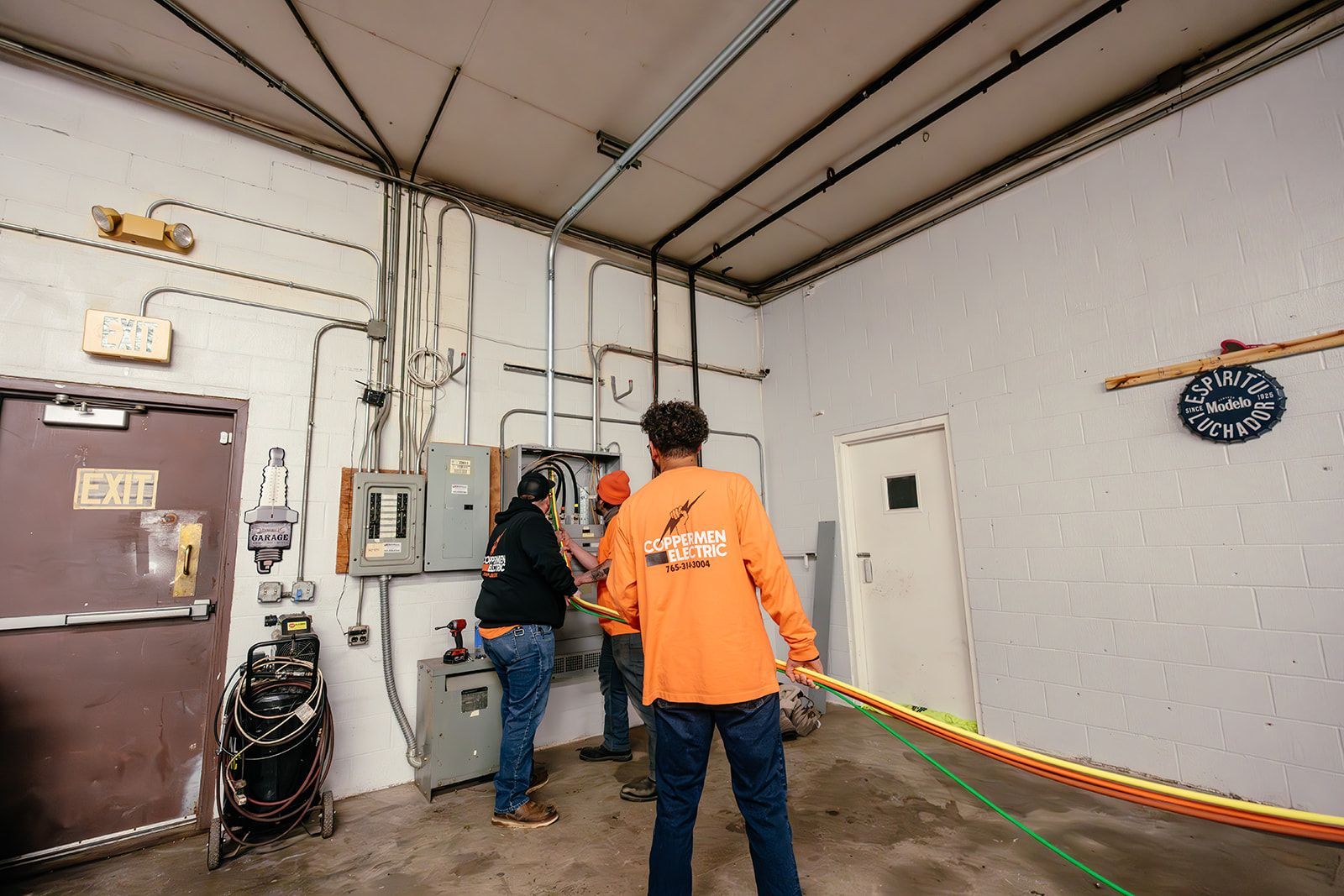 Three people in work clothes collaborate on electrical wiring at a breaker box in an industrial building.