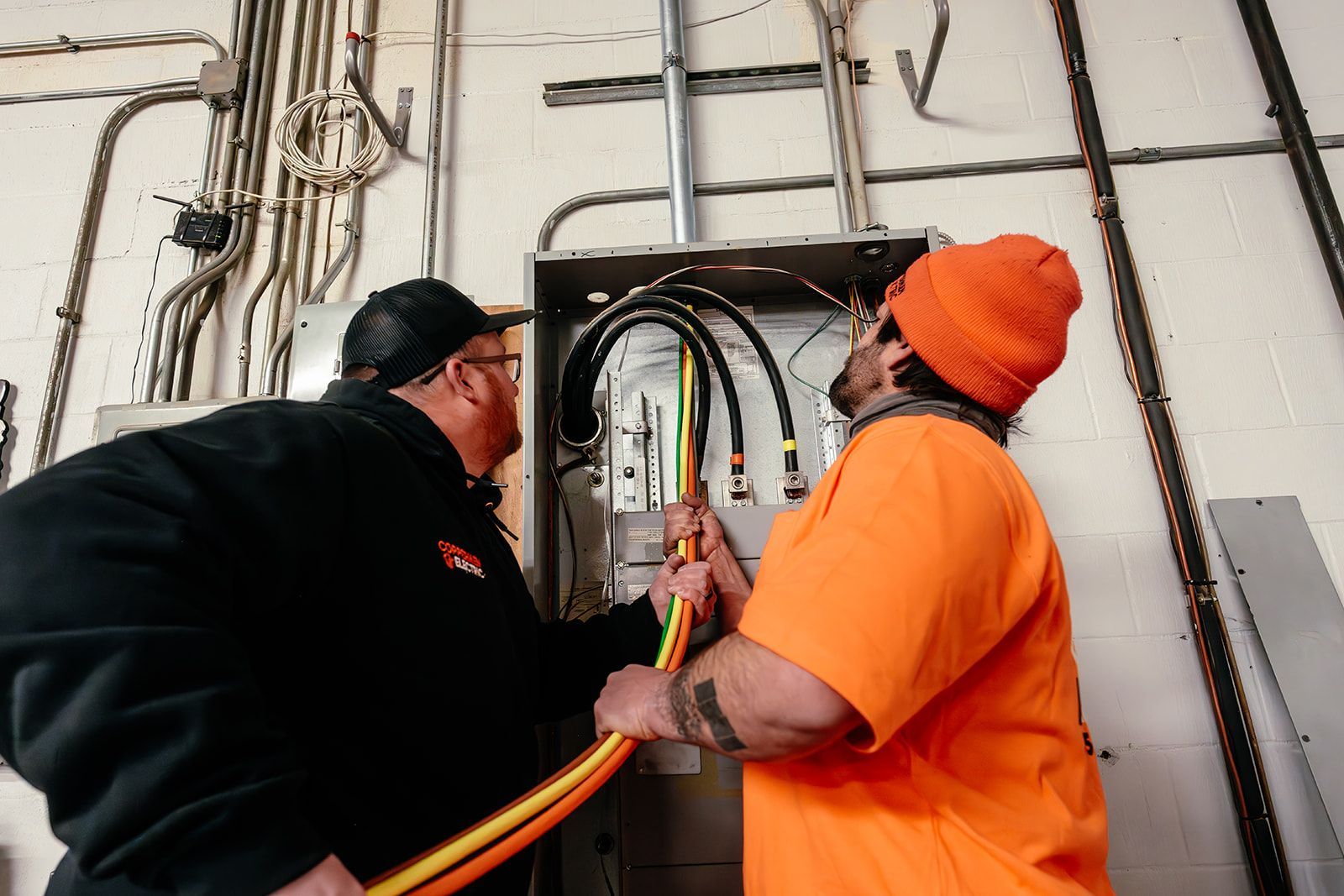 Two workers in high-visibility orange and black gear guide thick electrical cables into an open wall-mounted conduit box.