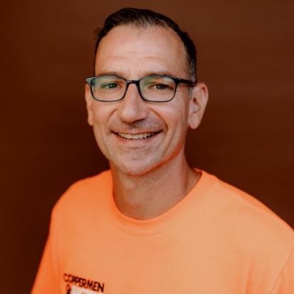 A smiling person wearing glasses and an orange t-shirt with a logo, set against a solid brown background.