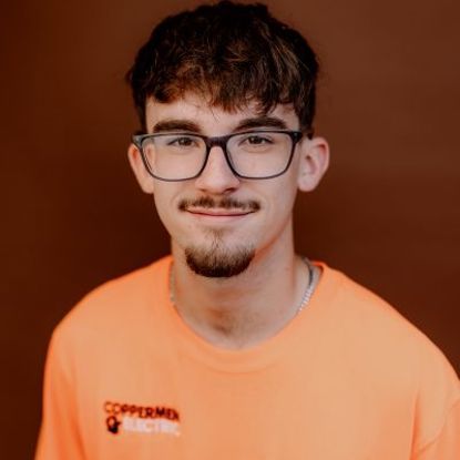 A young person wearing glasses and an orange Coppermine Electric t-shirt against a plain brown background.