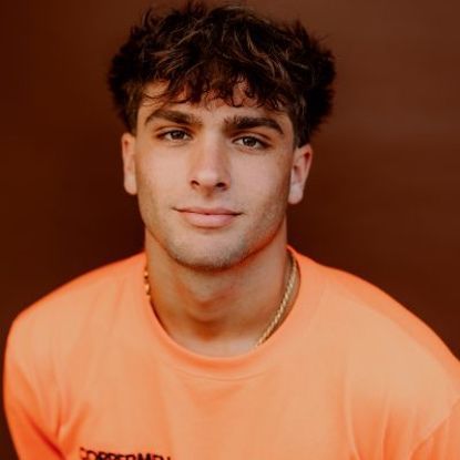 A person with dark, wavy hair wearing an orange shirt against a solid brown background, looking directly at the camera.