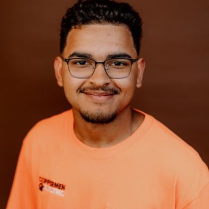 A person wearing a light orange t-shirt with a company logo on the chest, posing against a solid brown background.
