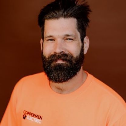 A person with dark hair and a beard, wearing an orange Coppermen Electric shirt against a solid brown background.