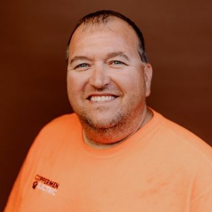 Smiling person wearing an orange shirt with a Coppermen Electric logo against a solid brown background.