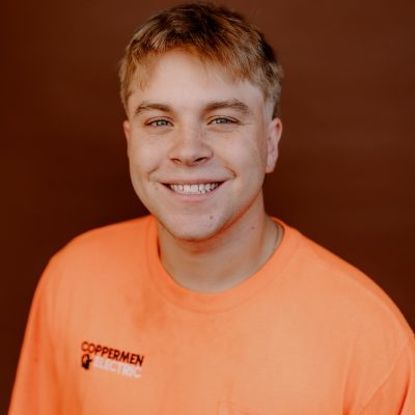 Smiling person wearing a bright orange Coppermen Electric T-shirt against a brown background.