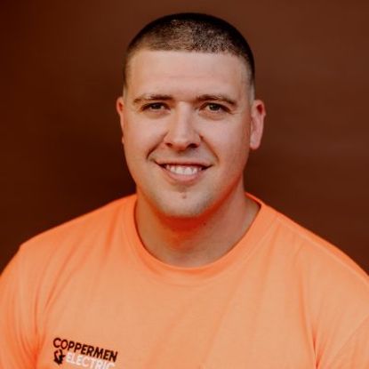 A smiling person wears an orange t-shirt with a Coppermin Electric logo against a solid brown background.