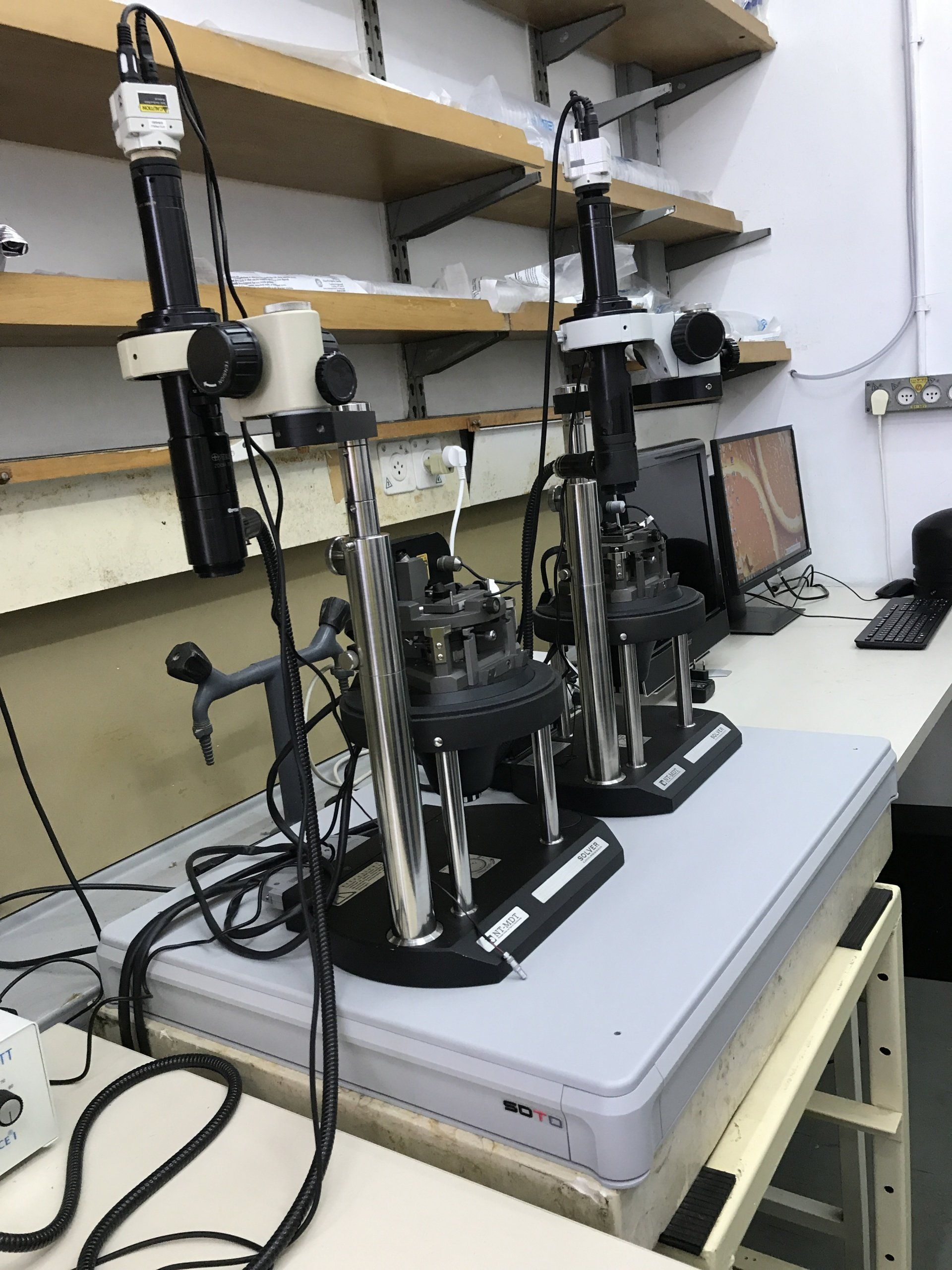Laboratory setup with two precision testing devices mounted on vibration isolation platform, next to workstation with monitor