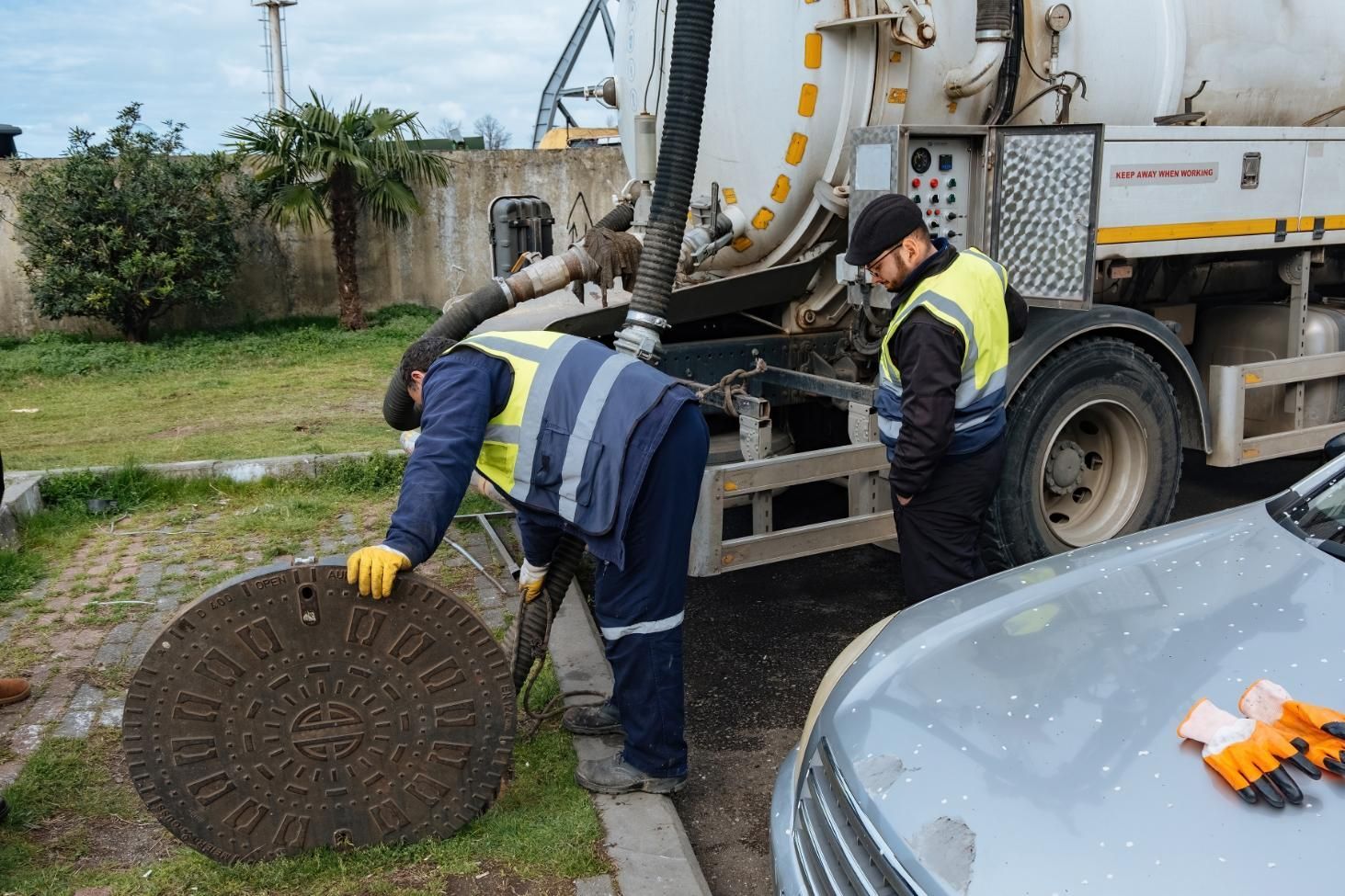 Two workers in reflective vests lift a manhole cover beside a utility truck on a grassy roadside.
