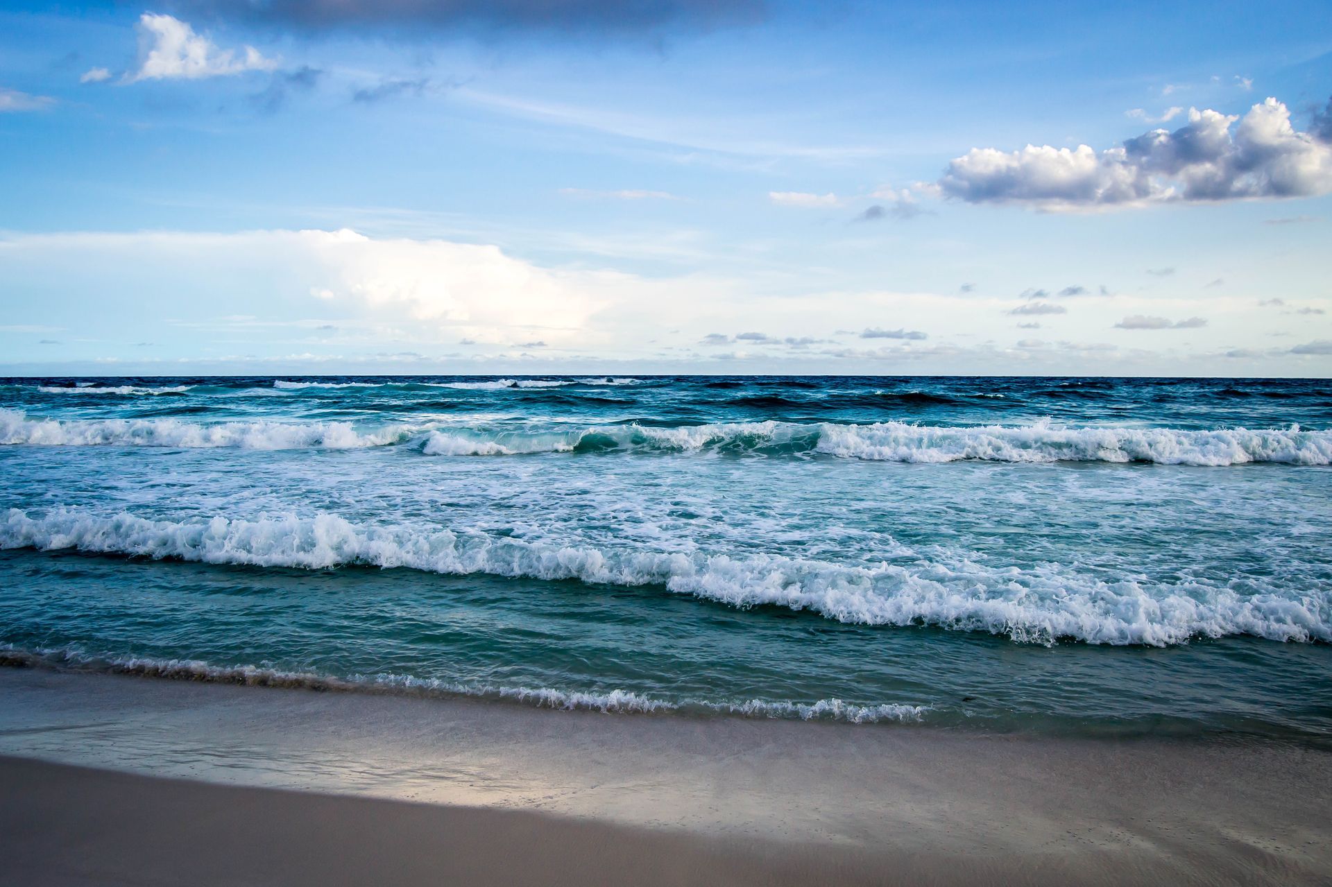 Waves rolling onto a sandy beach under a blue sky with white clouds.