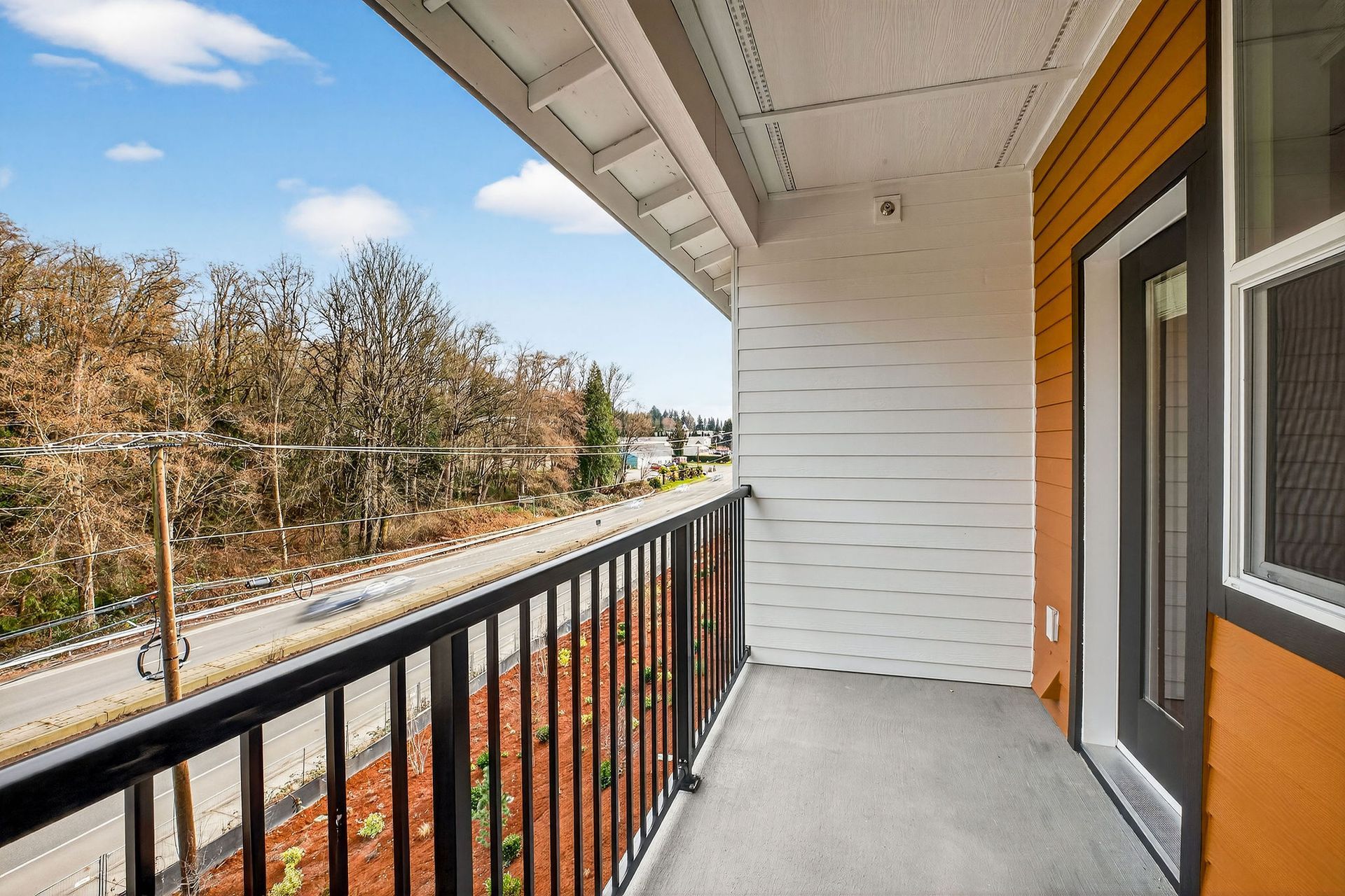 Balcony with black railing overlooking a road and trees, with orange siding and a door.