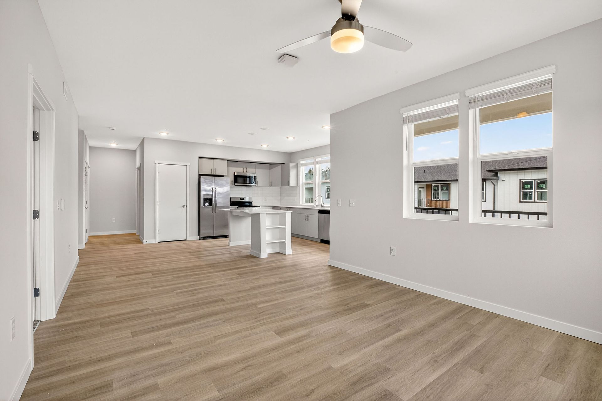 Empty modern living room with kitchen in the background, hardwood floors, large windows.