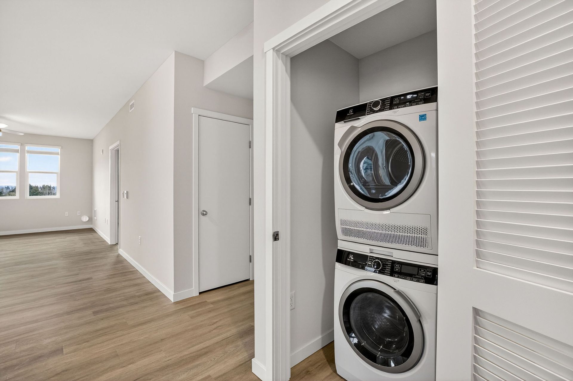 Stackable washer and dryer in a small closet, next to a hallway with doors and a window.