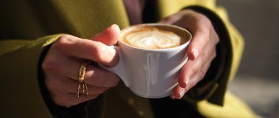 Hands holding a white coffee cup with latte art, person wearing a gold ring and a yellow jacket.