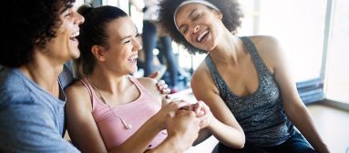 Three smiling people in workout clothes laugh together indoors.
