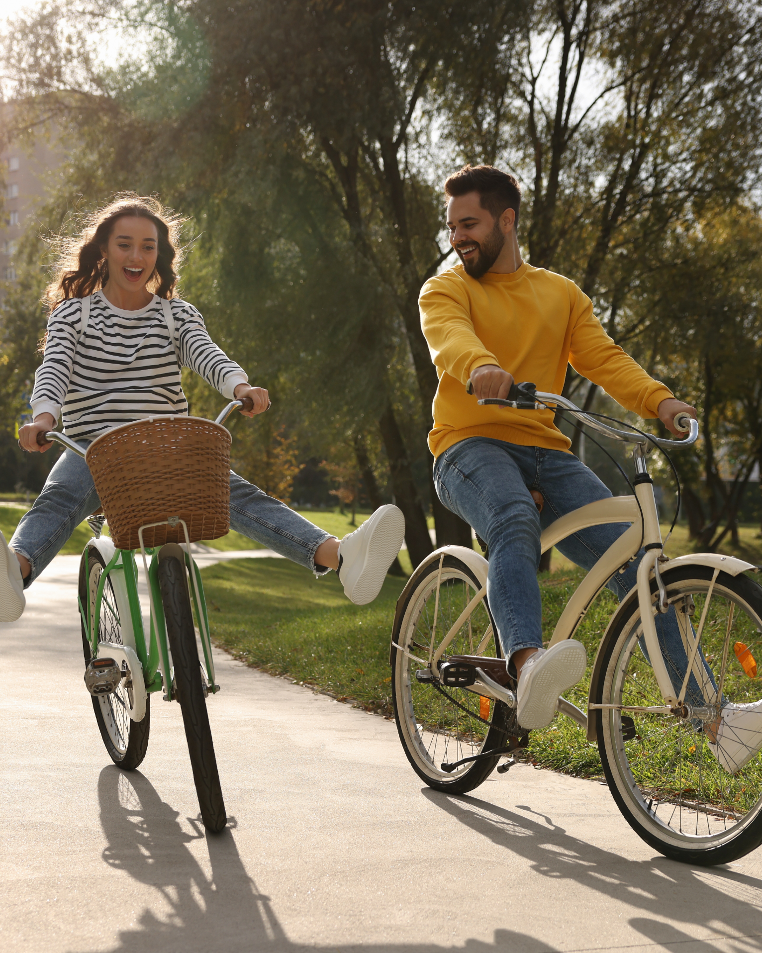 Two people ride bikes on a paved path in a park. One raises her legs and smiles. The other smiles.
