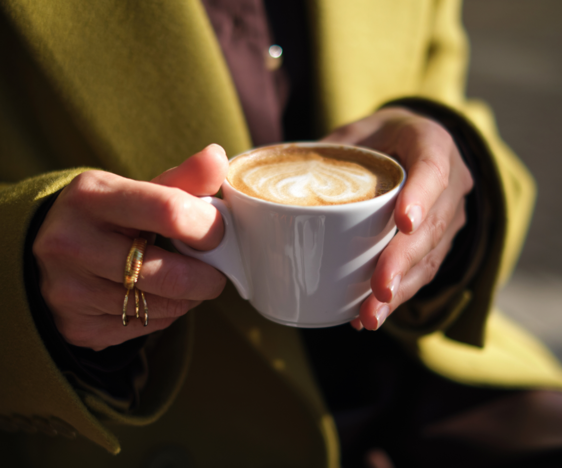 Hands holding a white cup of cappuccino with latte art, wearing a gold ring, with a yellow coat.