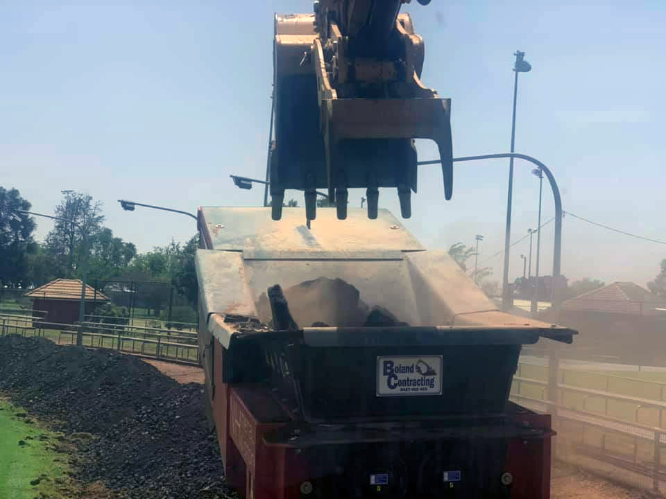 An Excavator Loading Dark Material Into a Truck at an Outdoor Construction Site — Boland Contracting In Wellington, NSW