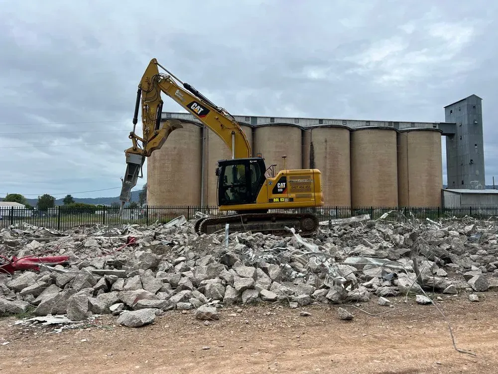 Yellow Excavator Demolishing Concrete Debris Near Silos on a Cloudy Day — Boland Contracting In Wellington, NSW