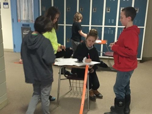 A group of children are standing around a desk in front of lockers