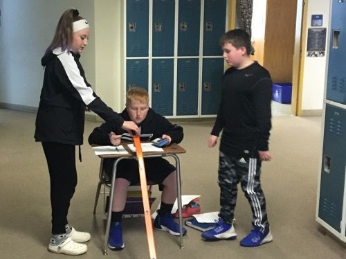 A boy is sitting at a desk while a girl stands next to him