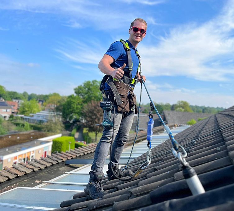 Worker in safety gear on a sloped roof, inspecting shingles under a blue sky.