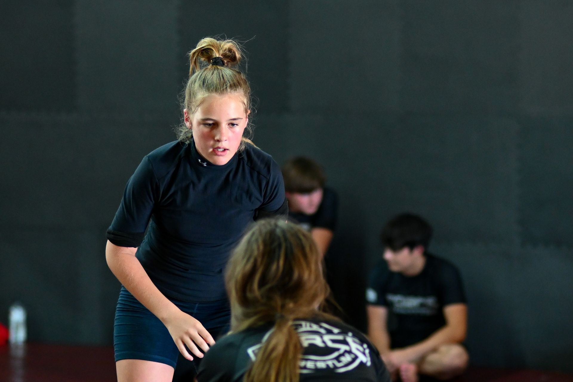 Girl in black grappling gear facing another person, in a gym. Other people are in the background.