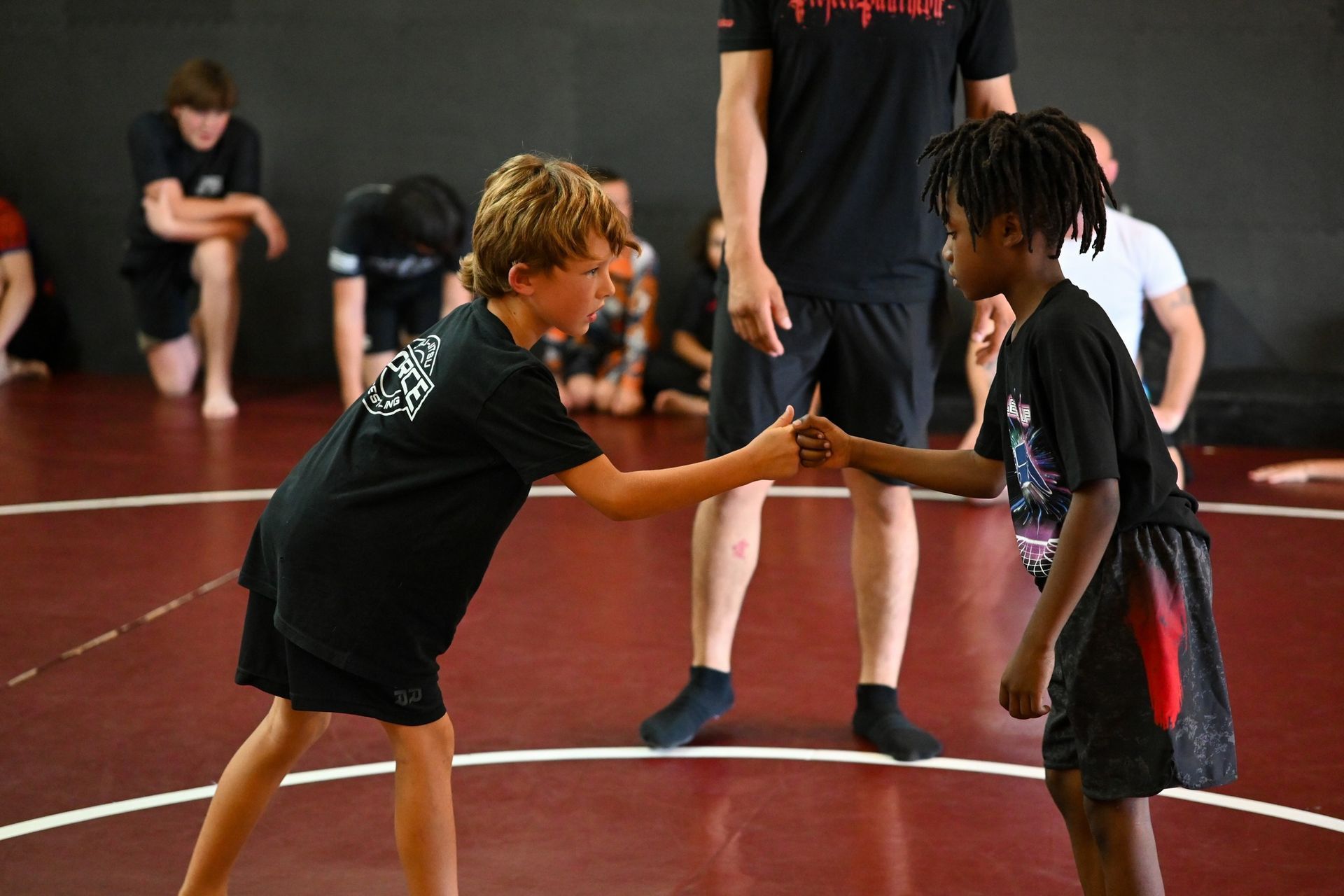 Two kids in wrestling gear bump fists on a wrestling mat. Other kids and a coach are in the background.