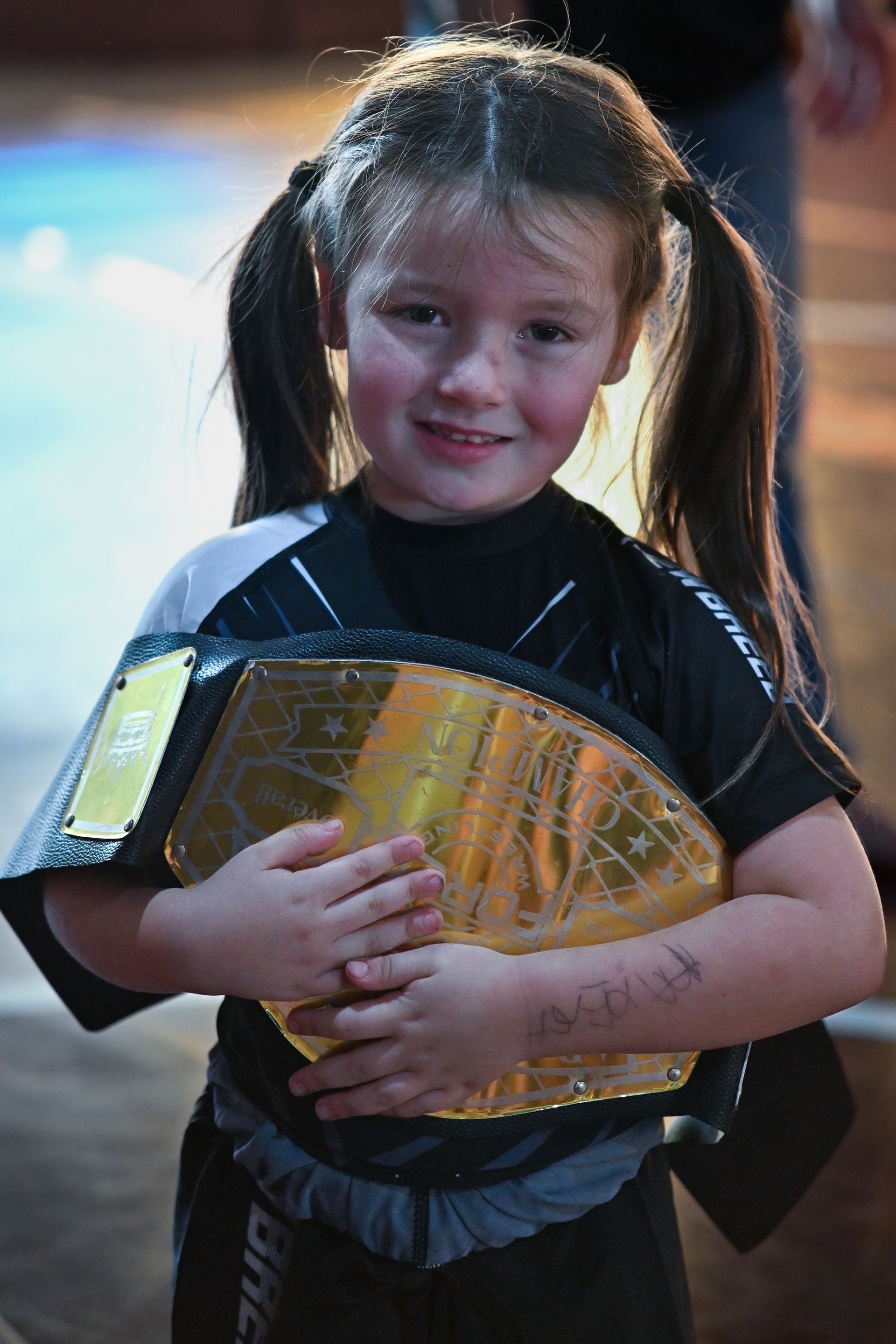 Girl in a martial arts uniform holding a championship belt, smiling at the camera.