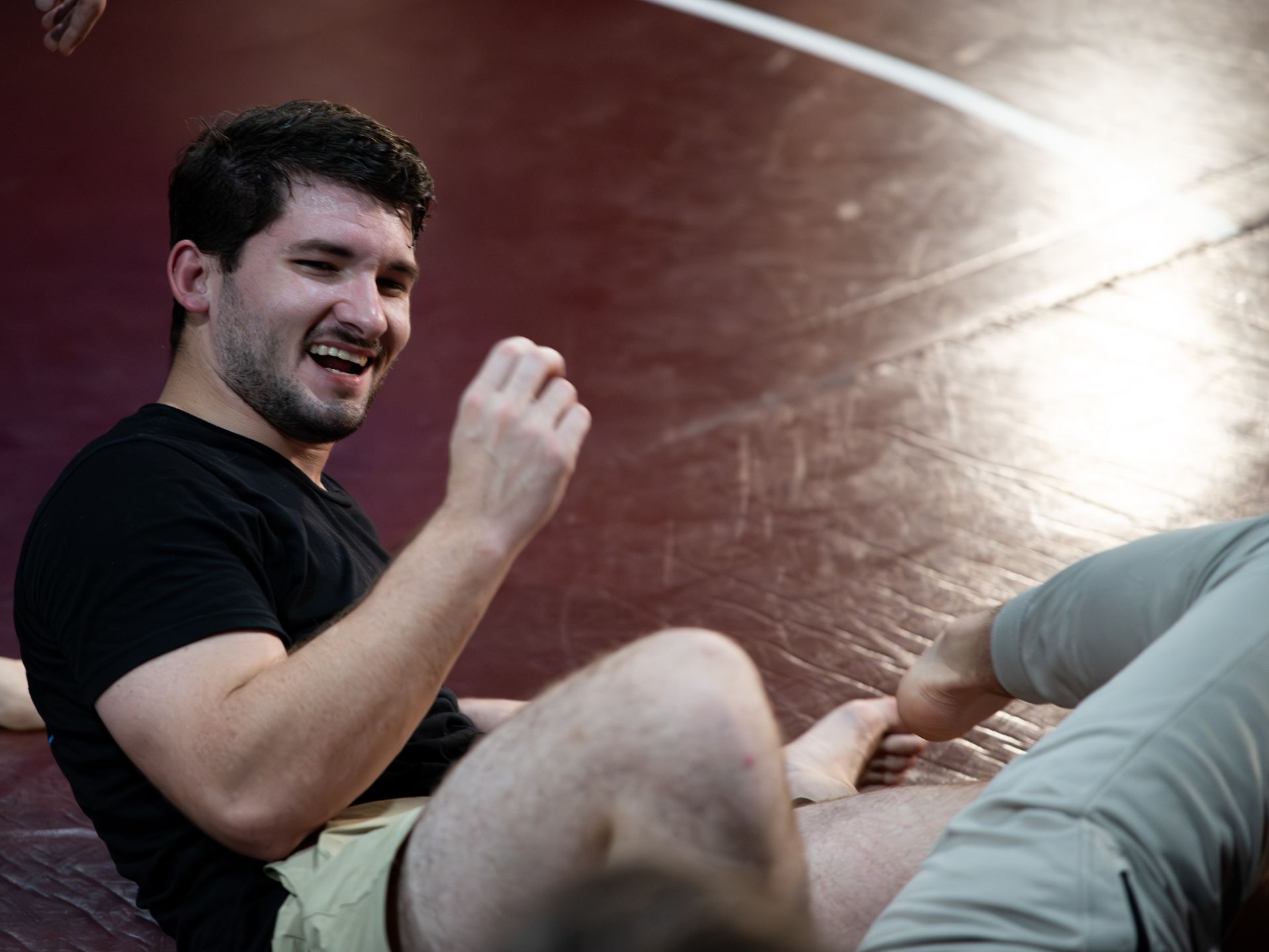 Man on wrestling mat, smiling. Arm raised, hand open. Other person's leg in foreground.