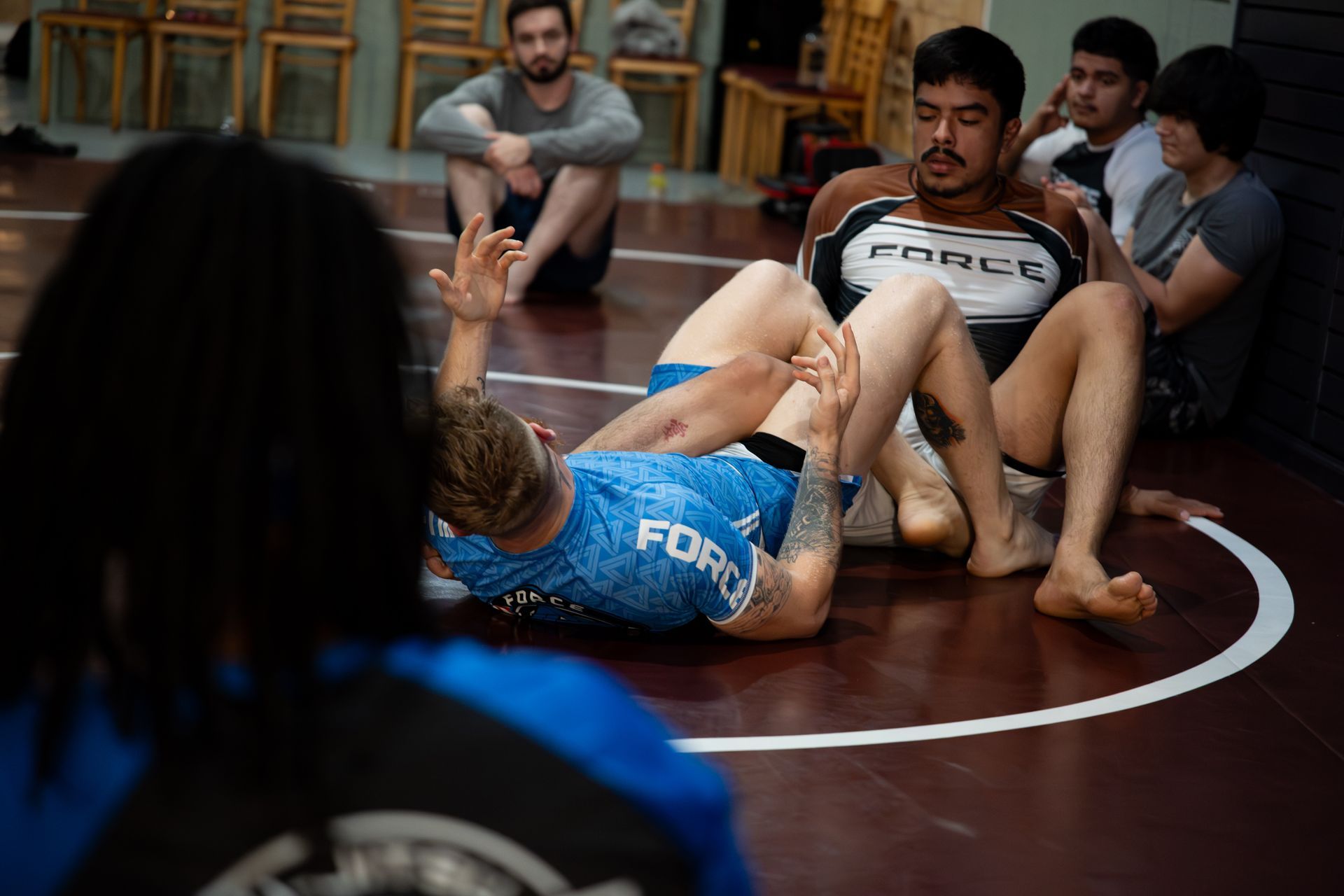 Two men in wrestling session on mat, others observing. One in blue shirt is on bottom, applying hold.
