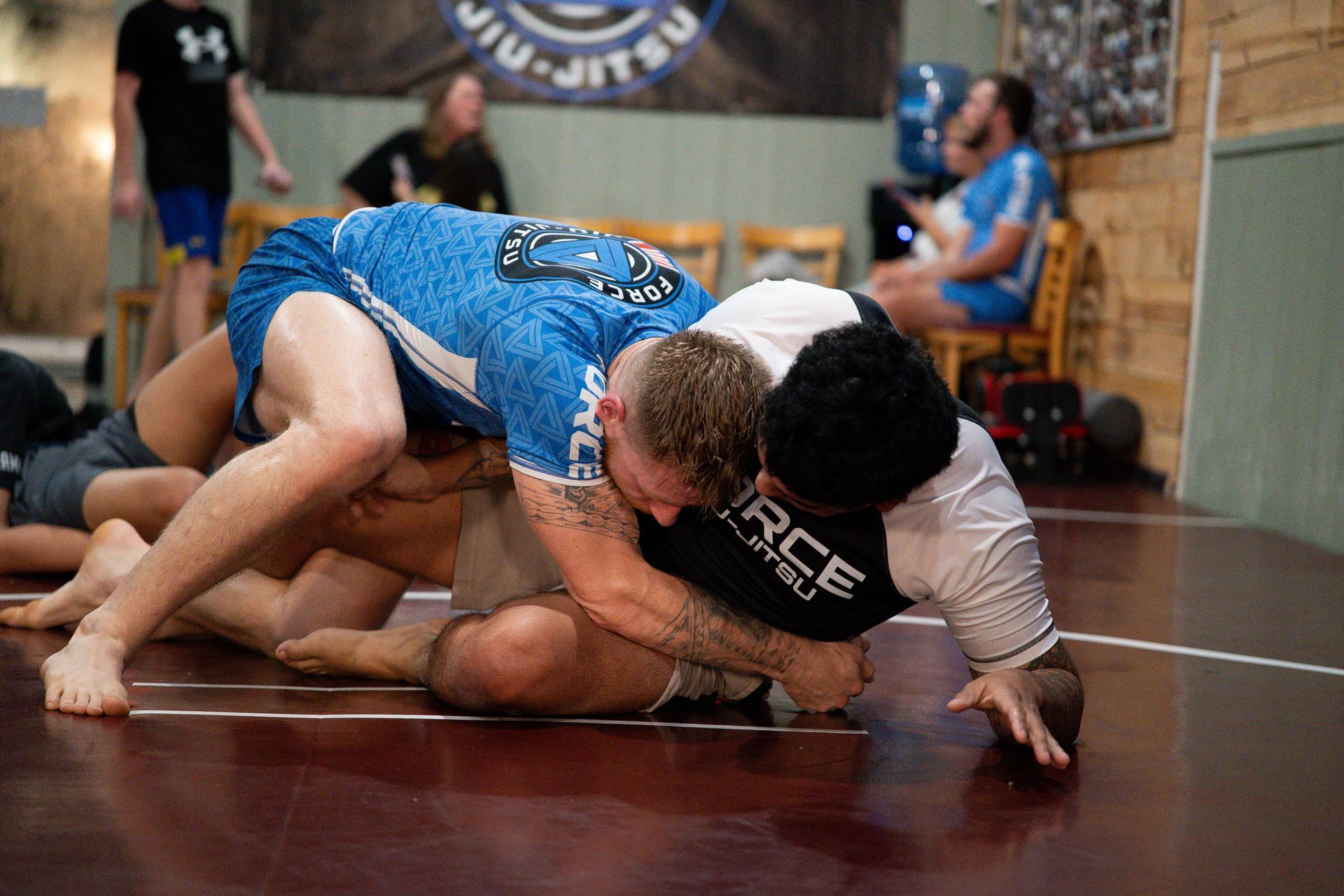 Two men grappling on a mat during a Jiu-Jitsu training session, others watching.